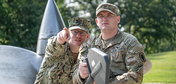 Image of a sailor and soldier, both in camo gear face towards the camera. Sailor pointing ahead, slightly towards the left while in a field with undescriptive communications gear is in the foreground.
