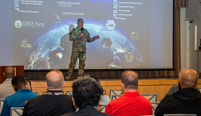 Brig. Gen. Cornell is on stage in the middle of the image while facing an audience. Eight members of the audience, in the foreground, have their backs to the camera. On a screen behind Cornell is a slide of the DISA NEXT Strategy.