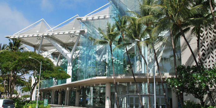 Photo of the Hawaii Convention Center. A building with triangular shapes and lots of glass with palm trees on the sidewalk.