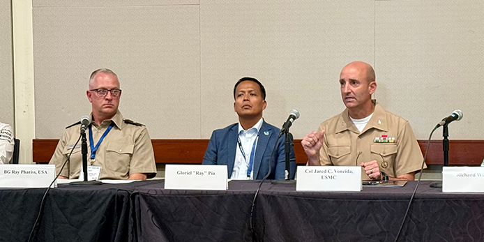 Photo of Army Brigadier General Ray Phariss, Gloriel "Ray" Pia and Marine Corps. Colonel Jared C. Voneida, with Voneida speaking sitting behind a long table with their names displayed on name tents  and microphones  in front of them.