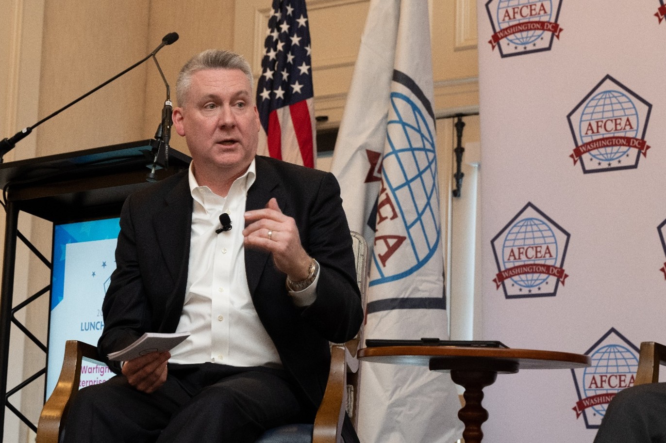 Jason Martin is pictured on the left side looking towards the right while sitting on an wooden chair. He's wearing a suit, collard shirt with the top button open and no tie. He has a lapel mic clipped on his shirt. Behind him, from left to right, there is an empty lectern, the national ensign, an AFCEA flag and an AFCEA Washington D.C. backdrop. In his right hand he holds large notecards, and to his left there's a wooden table.