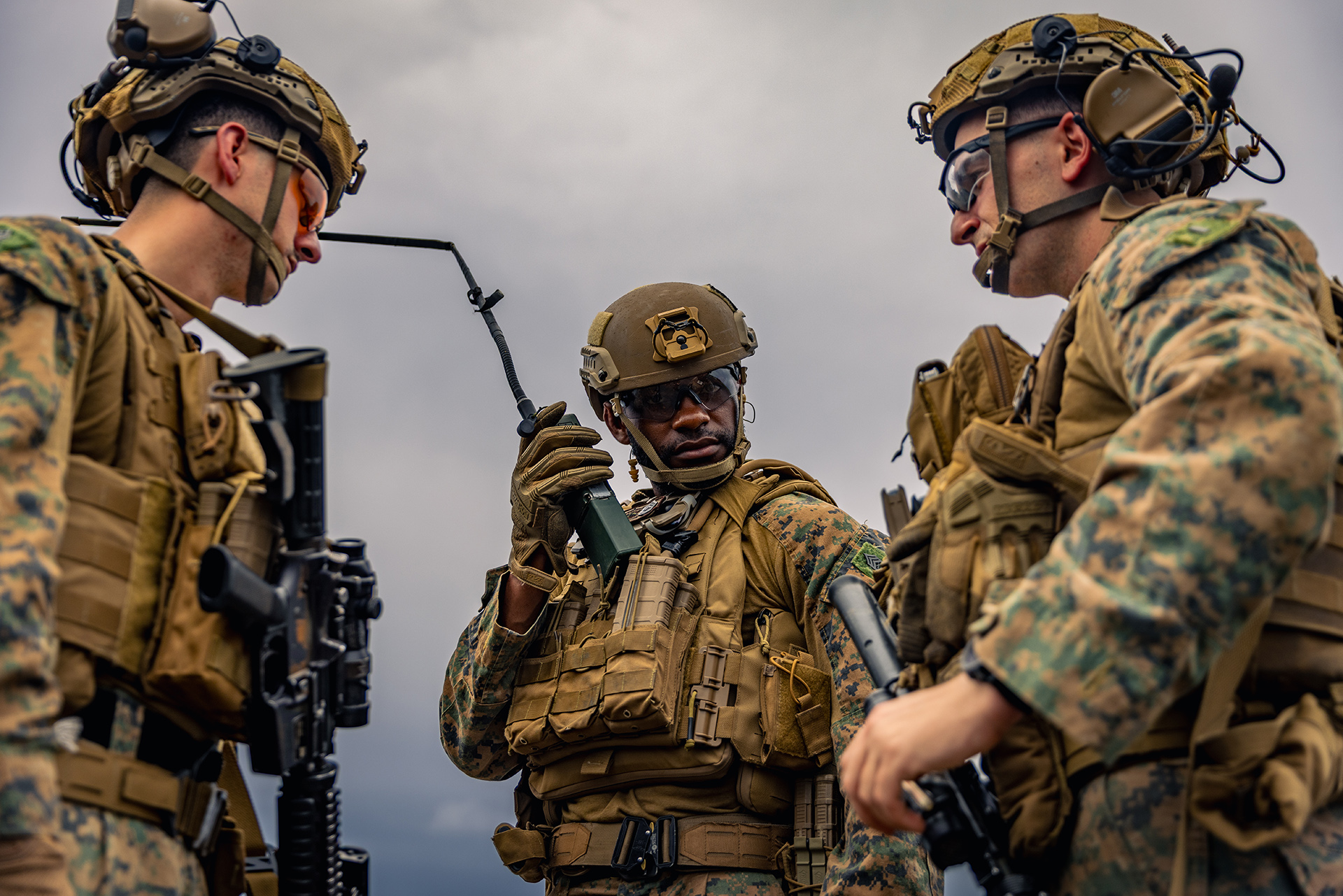 Photo of three Marines standing in full battle gear with the one in the middle facing the camera and the two others on each side facing eachother. The one in the center is holding up a radio near his right ear.