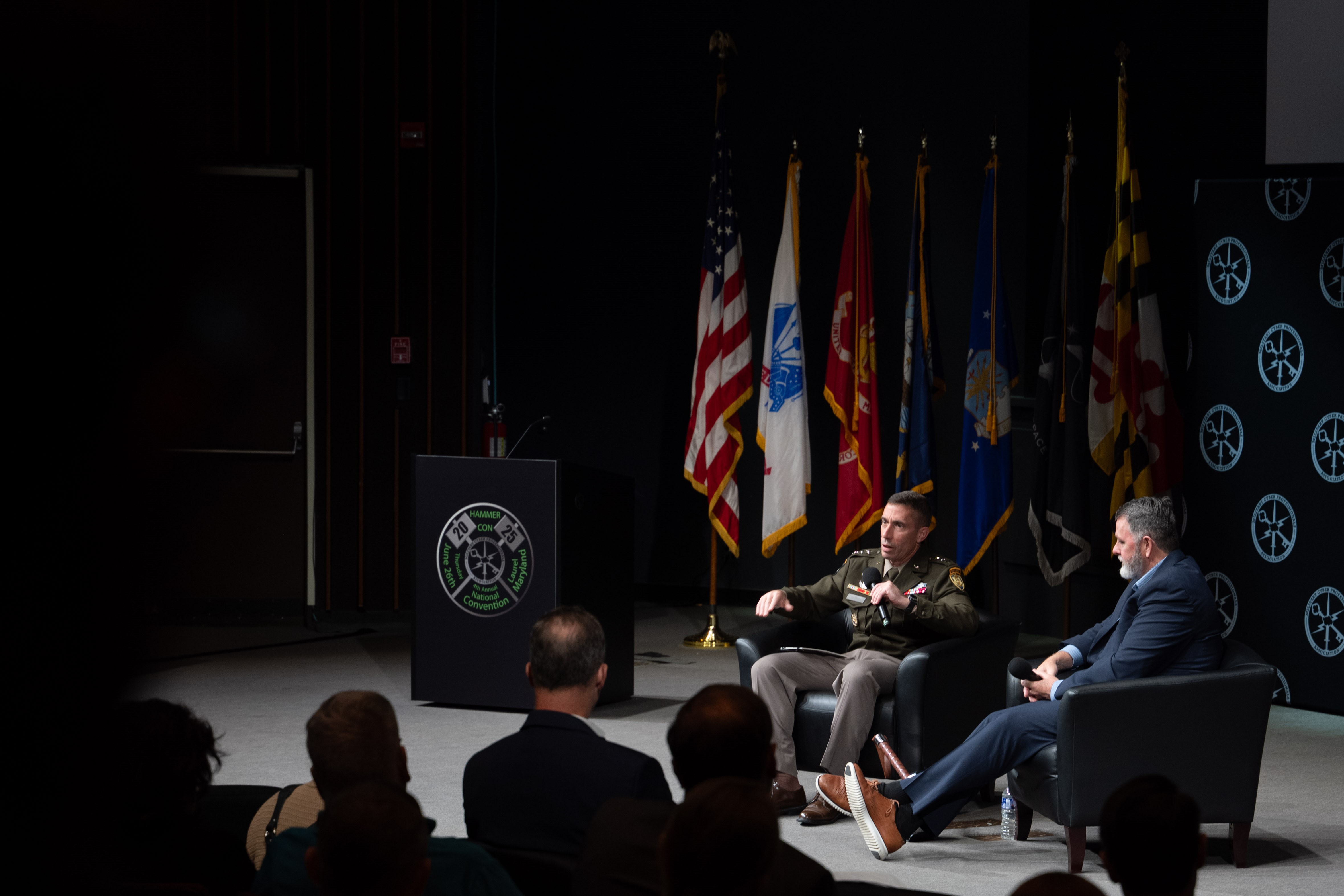 Photo of Lt. Gen. Stanton on stage with moderator Chris Cleary, near the lower right corner, facing to the left. The audience is in the lower left corner foreground facing away from the camera. Behind Stanton and Cleary is the national ensign, the flags of the services and the state flag of Maryland.