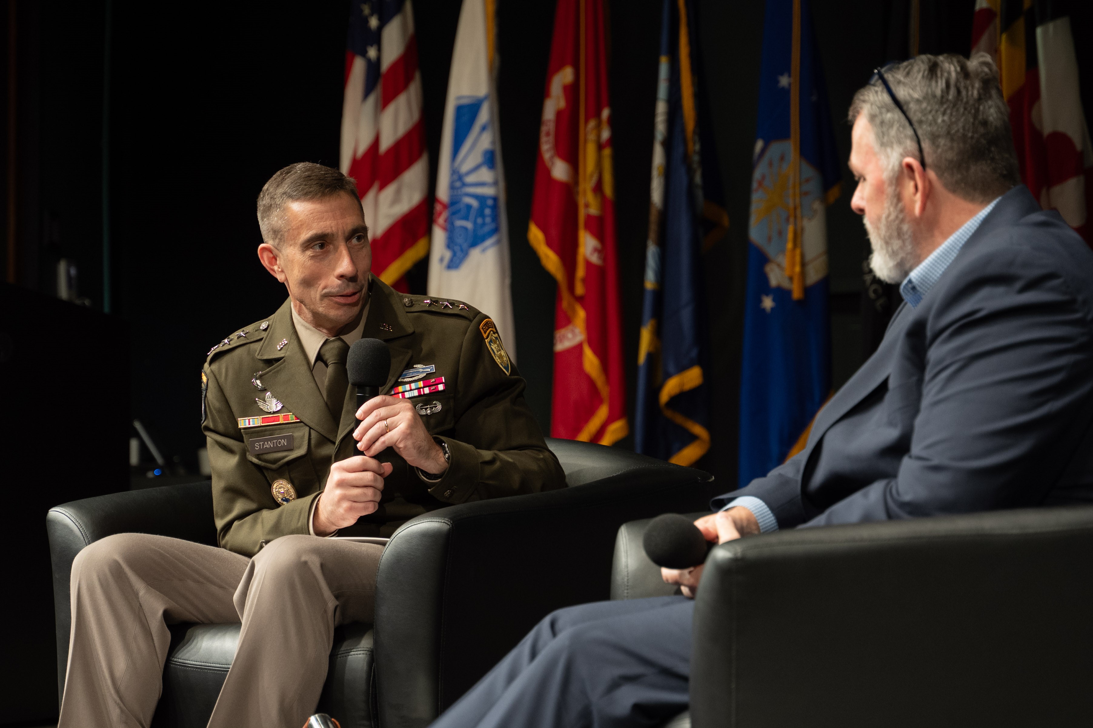 Photo of Lt. Gen. Stanton on stage looking towards the right at moderator Chris Cleary, who is facing Stanton and away from the camera. Behind Stanton and Cleary is the national ensign, the flags of the military services.